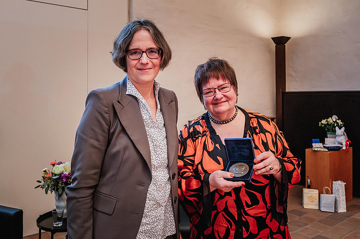 Verabschiedung und Auszeichnung von Dr.in Ursula Fuhrich-Grubert Dr.in Ursula Fuhrich-Grubert hält die Humboldt-Medaille in die Kamera und lächelt. Sie trögt eine orange-schwarz gemusterte Jacke und ein schwarzes Shirt. Julia von Blumentahl steht neben ihr und trägt einen taupe-farbenen Blazer und eine weiß-schwarz gemusterte Bluse.